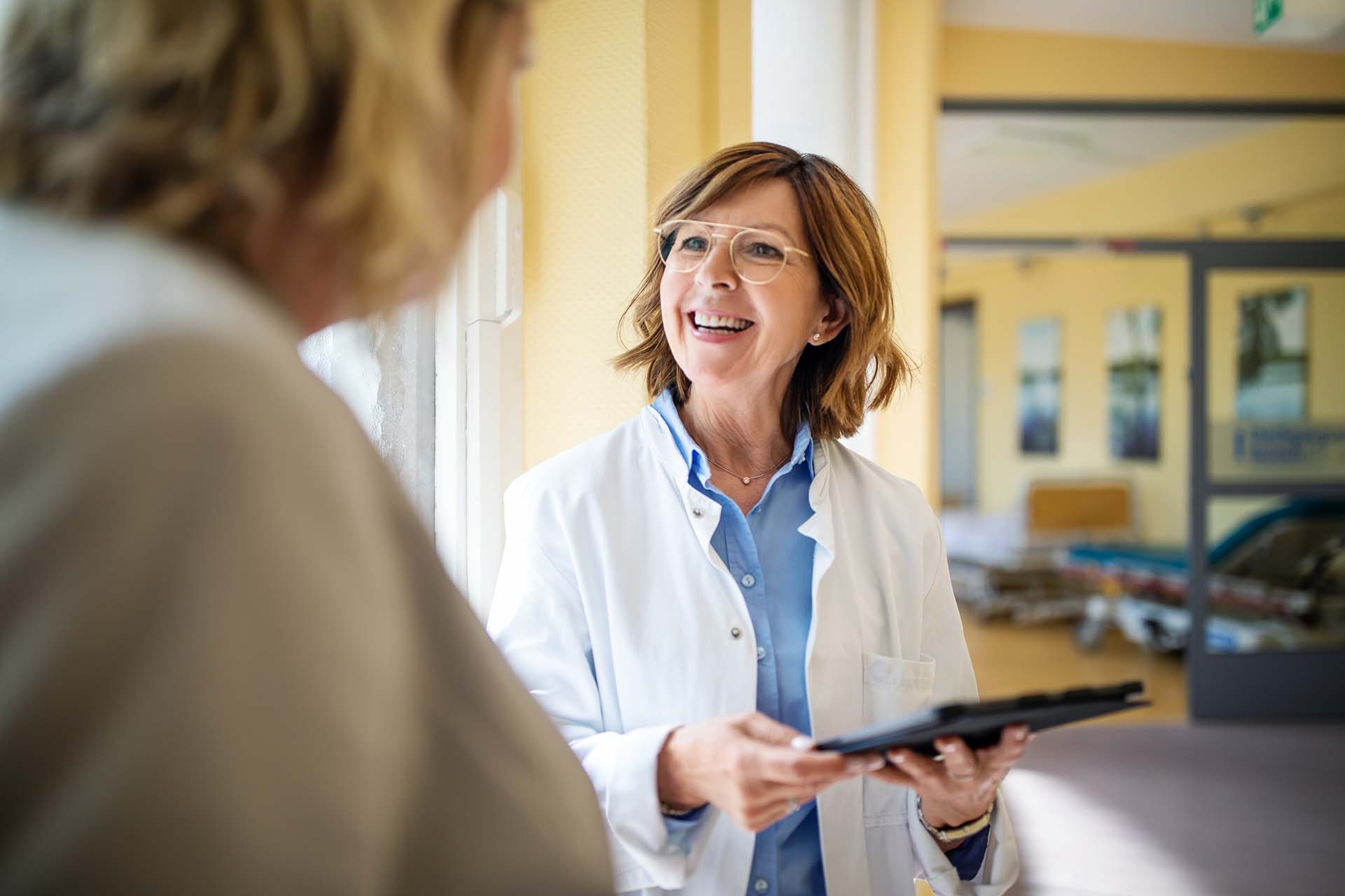 Shot of a smiling senior doctor with a digital tablet talking to a female patient at hospital. Medical professional discussing with woman at nursing home.