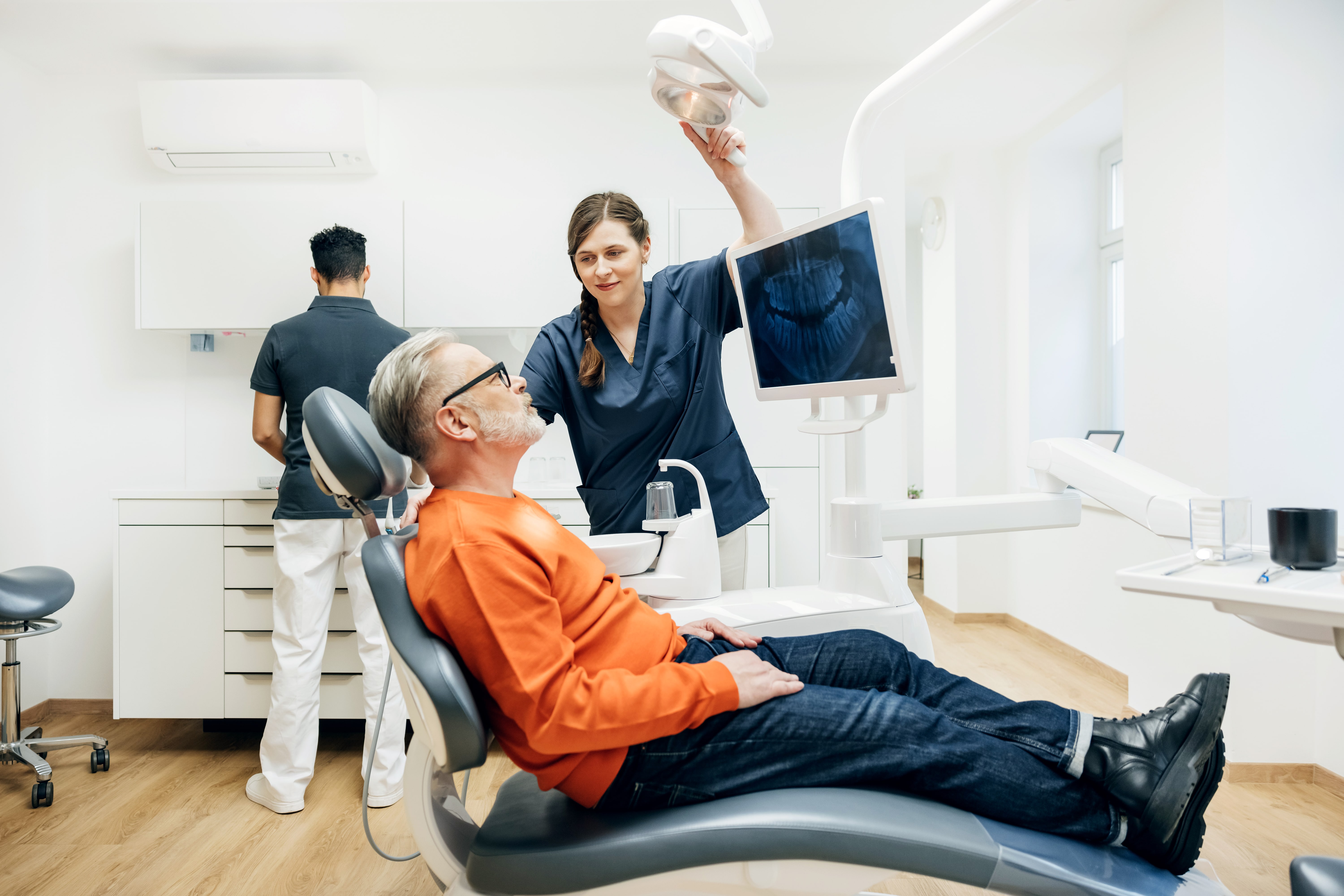A dental assistant adjusting an overhead light during a check up on a patient sitting back in a mechanical chair while the orthodontist prepares for a procedure.